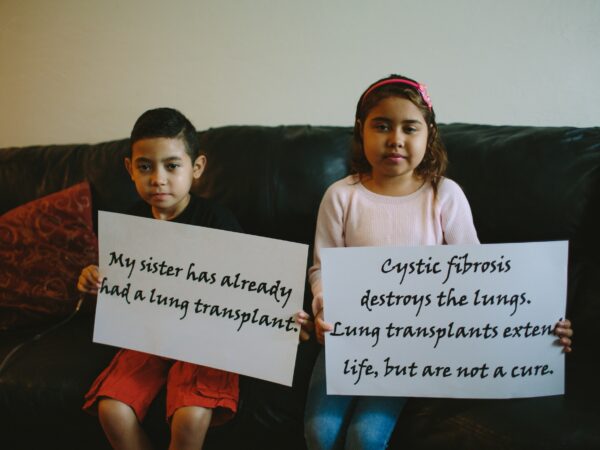 a young boy and girl sitting side by side on a couch holding signs that read; "my sister has already had a transplant" and cystic fibrosis destroys the lungs. Lung transplants extend life but are not a cure"