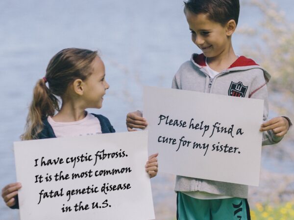 HannaCFMostCommonGenetic Cropped Image of 2 children, a boy and girl holding signs and looking at each other, the signs say "i have cystic fibrosis. its th emost common genetic disease in the us." and the othetr says please help find a cure for my sister"